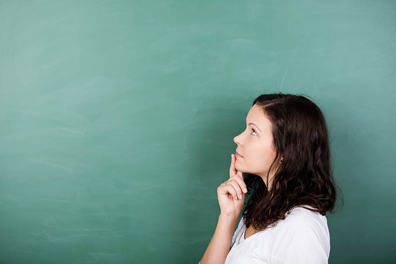 Learning Center 11 A woman with long brown hair, wearing a white shirt, stands in profile against a green chalkboard, looking thoughtful with her finger resting on her chin.